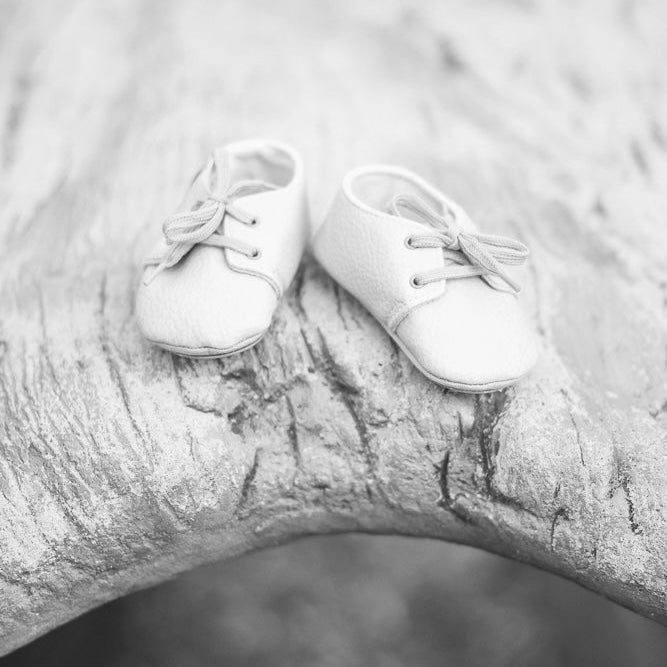 A pair of white leather baby booties with laces, resting on a textured, curved piece of driftwood in a black and white photograph, preparing for Grace that was the inspiration for EverGrace Coffee.