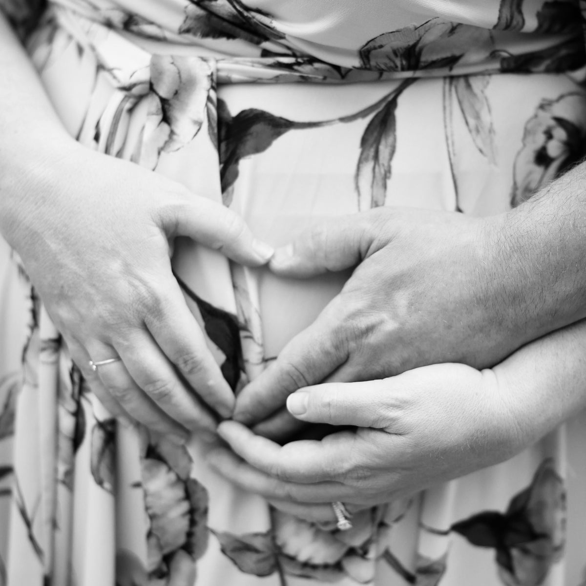 Black and white close-up of two people's hands gently holding a baby bump, with a floral-patterned dress visible in the background.