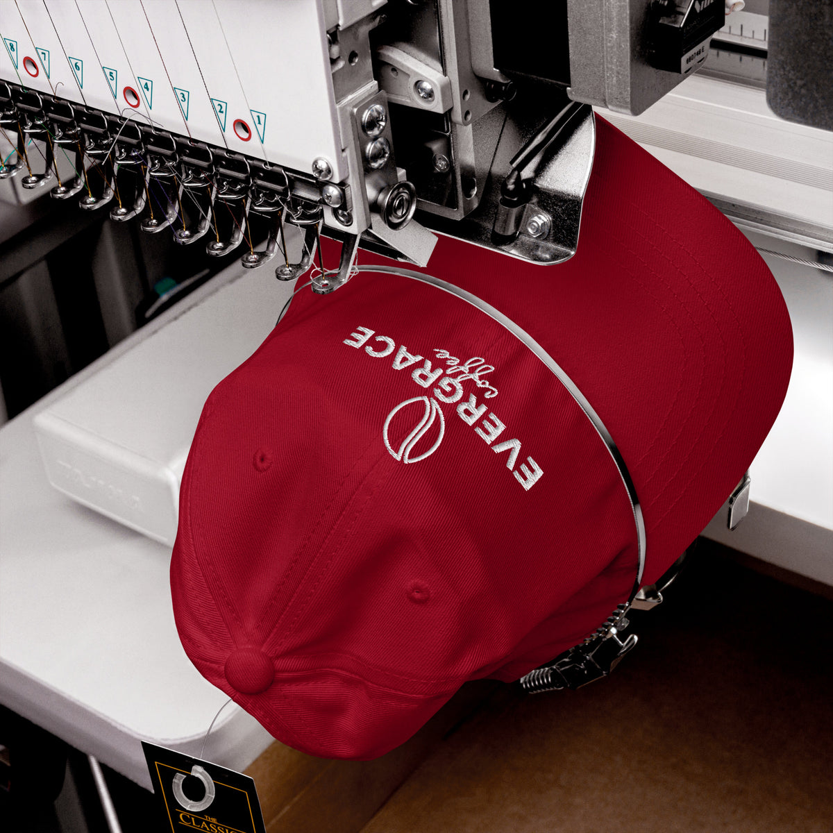 A red baseball cap with white embroidered text reading 'EverGrace Coffee' is being held by a metal hoop over an industrial embroidery machine.