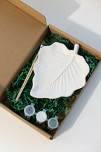 A plater leaf-shaped painting palette set inside an open brown cardboard box filled with green crinkle paper. The set includes a white leaf-shaped palette, a wooden stick, and three small paint cups with white lids.
