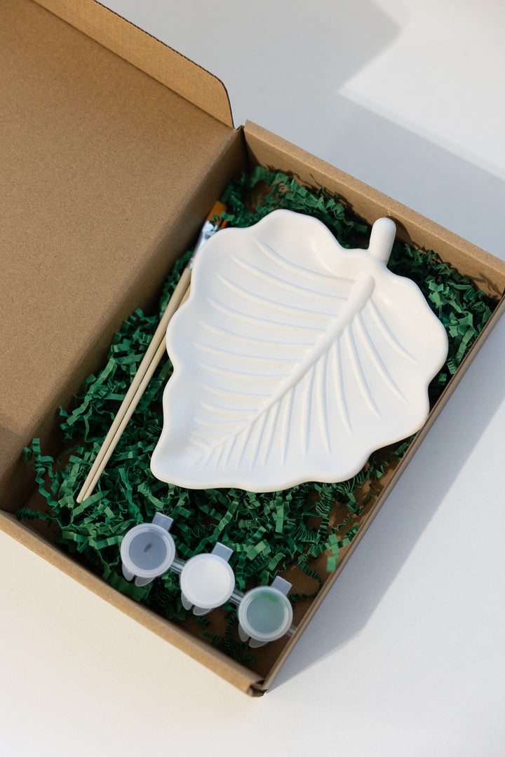 A plater leaf-shaped painting palette set inside an open brown cardboard box filled with green crinkle paper. The set includes a white leaf-shaped palette, a wooden stick, and three small paint cups with white lids.