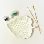 A plaster leaf-shaped paint palette with embossed leaf veins, shown on a white background with two wooden paintbrushes and small paint pots from EverGrace Coffee and Crafts