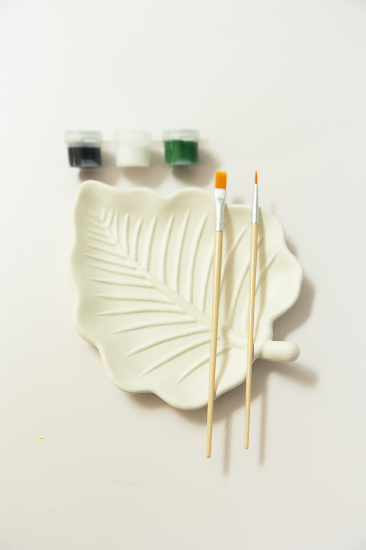 A plaster leaf-shaped dish in a warm off-white glaze, styled with two wooden paintbrushes and three small paint pots on a clean white background from EverGrace Coffee & Crafts.