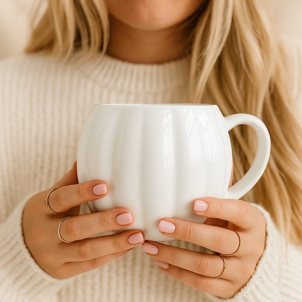 A close-up lifestyle shot of a woman in a cream knit sweater holding a white ribbed ceramic mug with both hands. She is wearing delicate gold rings.