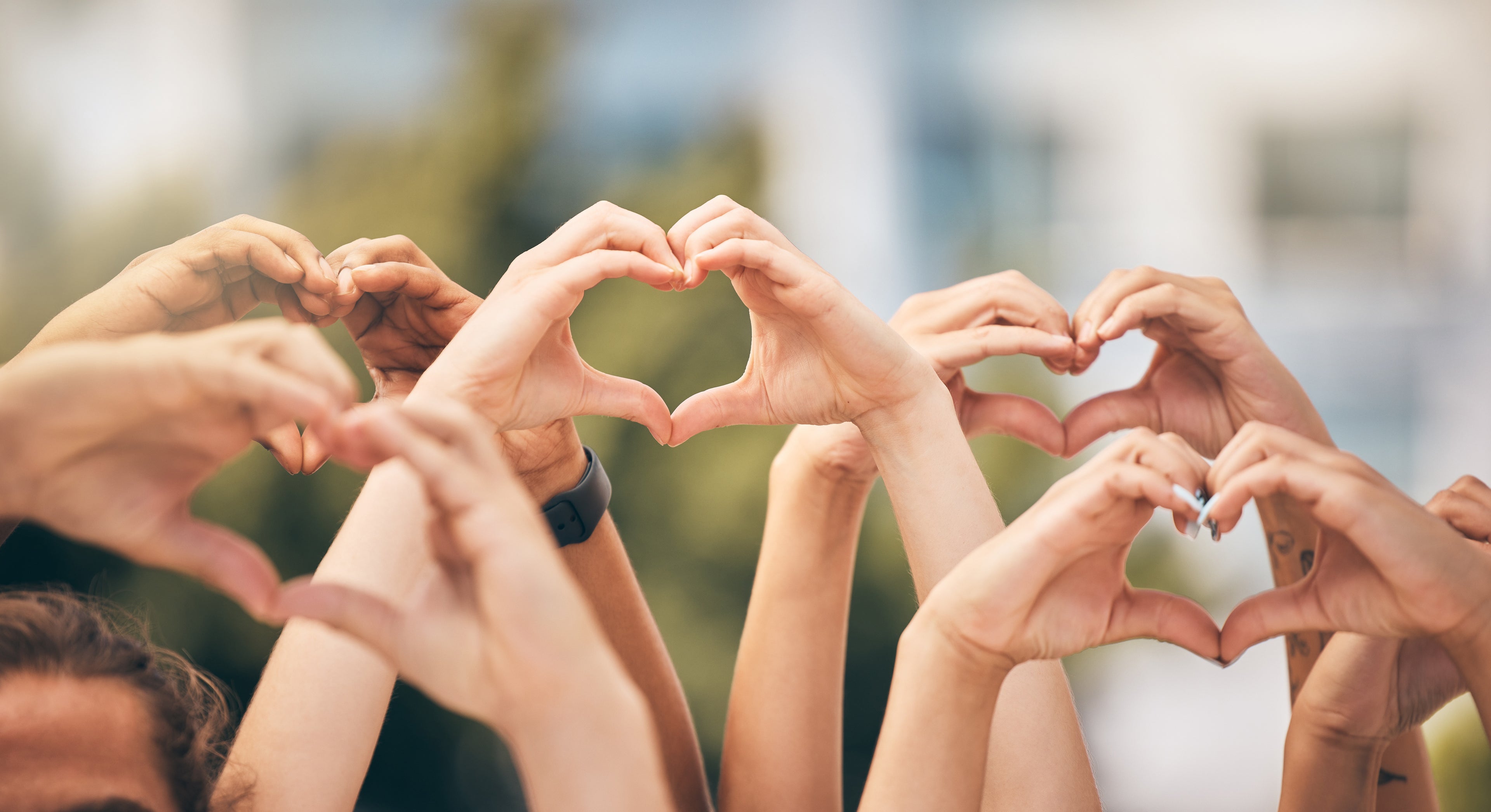 Multiple hands raised together to form heart shapes against a softly blurred outdoor background, showing shared graces with the EverGrace Giving Program.