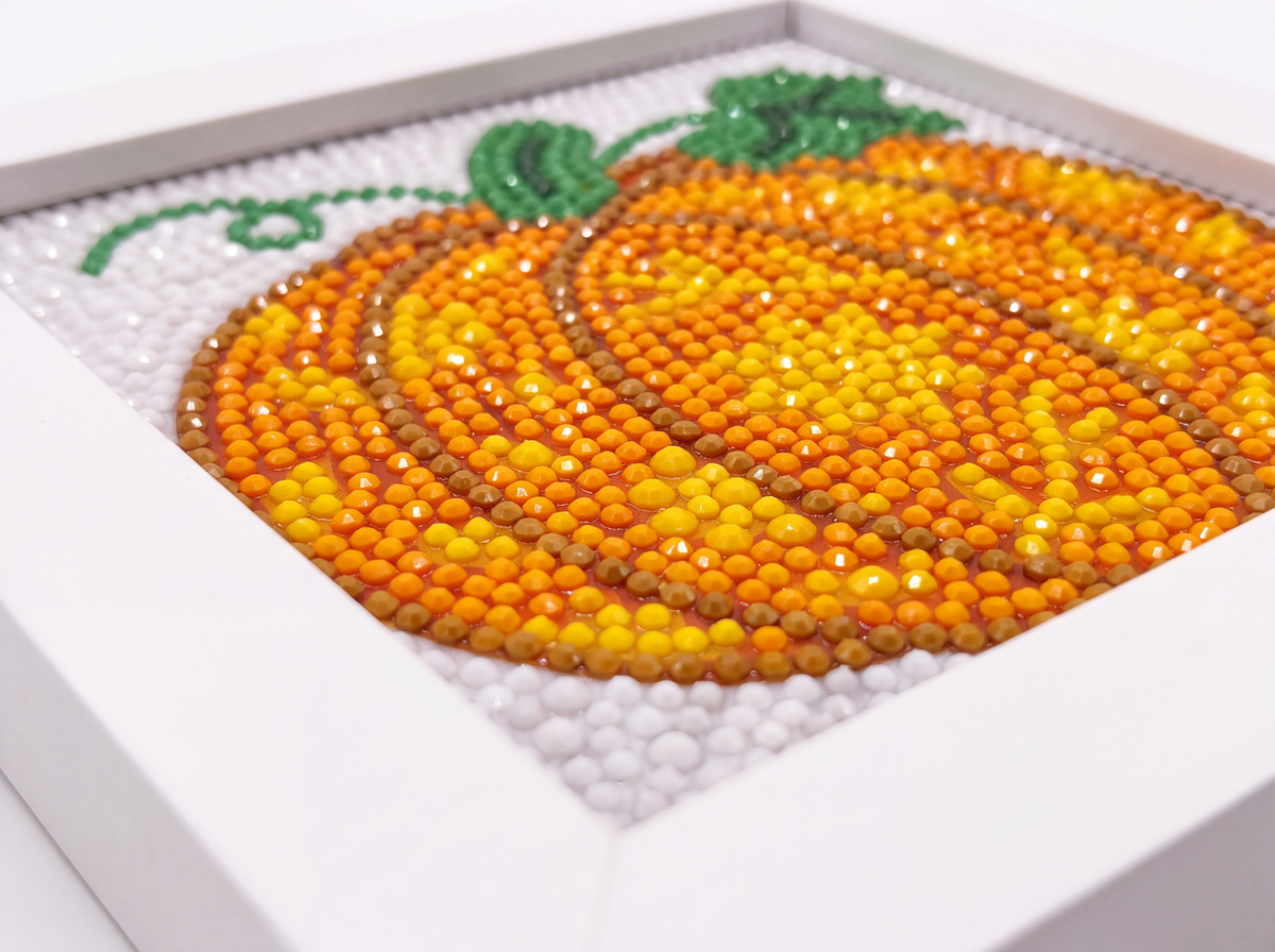 A diamond painting or bead art piece of a pumpkin in orange, yellow, and brown, with a green stem and leaves, set in a white frame.