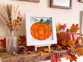 A framed diamond painting of an orange pumpkin with green leaves, displayed on a rustic wooden shelf in an autumn-themed setting with artificial leaves, a small pumpkin, and dried grass in a glass vase.
