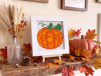 A framed diamond painting of an orange pumpkin with green leaves, displayed on a rustic wooden shelf in an autumn-themed setting with artificial leaves, a small pumpkin, and dried grass in a glass vase.